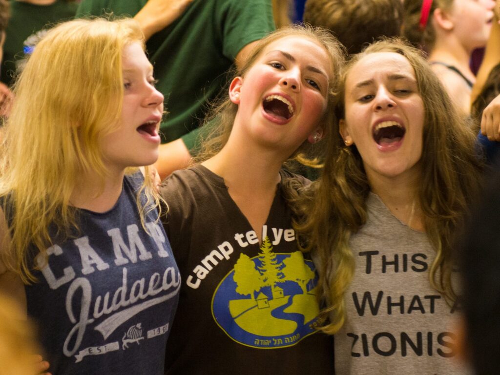 Campers arm in arm singing, one in a Camp Judaea shirt and one in a Camp Tel Yehudah shirt